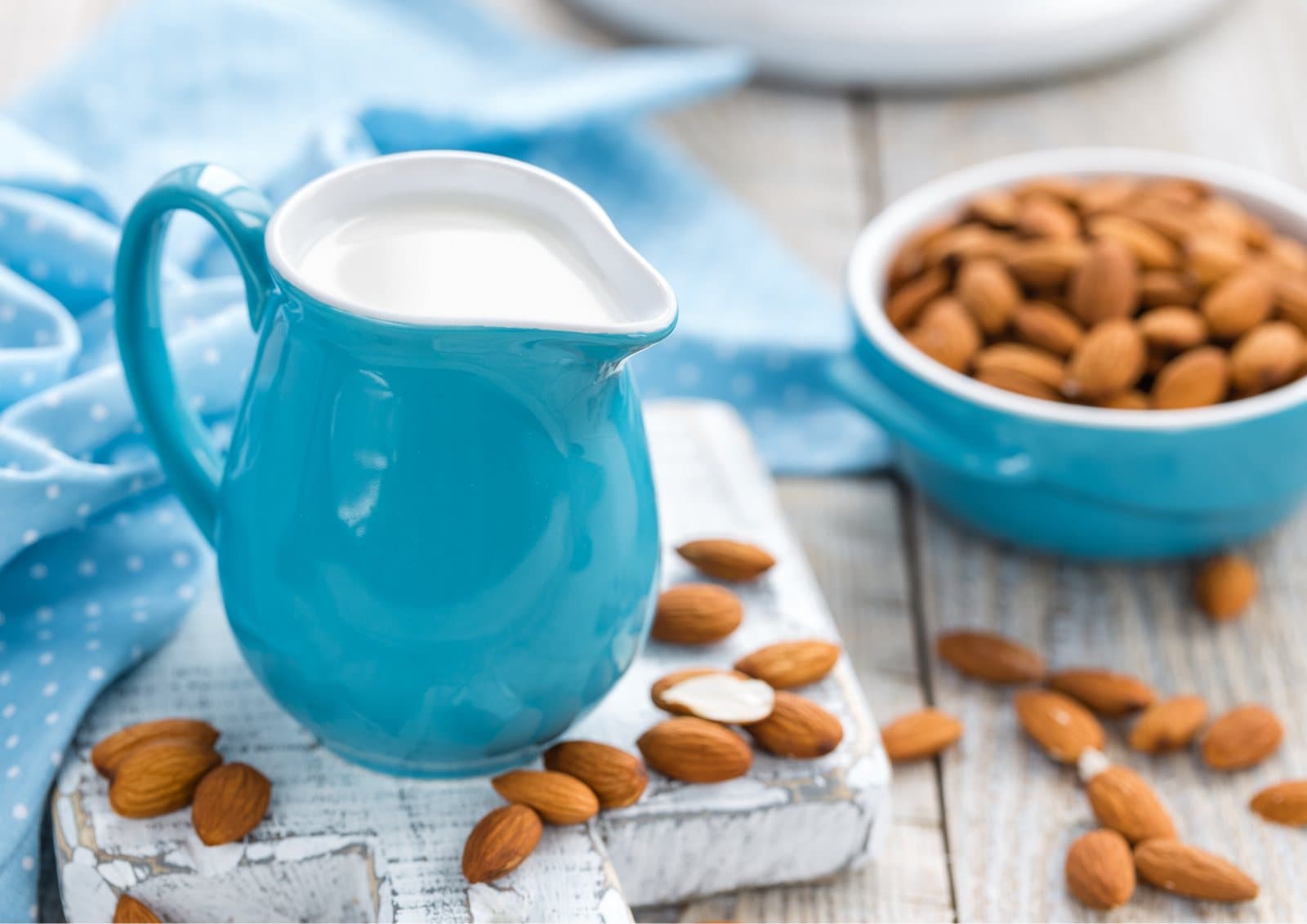 A blue jug of almond milk on a board beside a bowl of almonds.